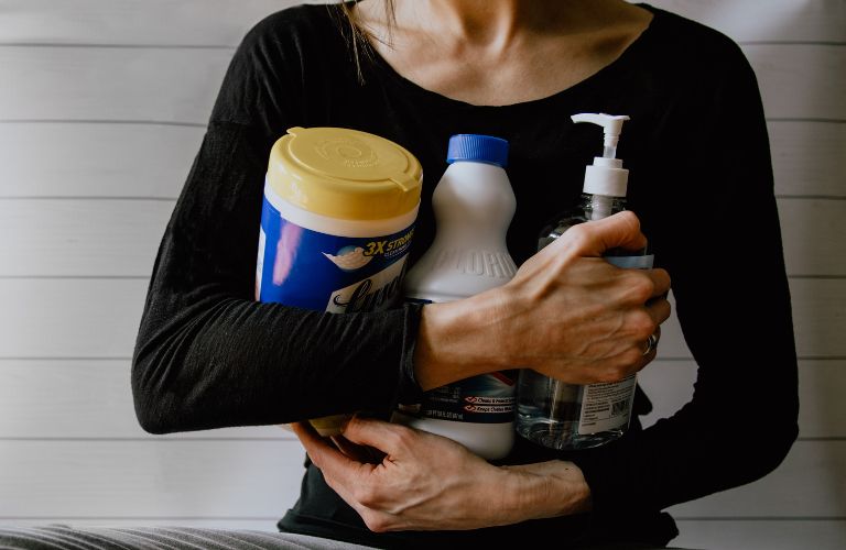 woman holding multiple cleaning soaps and detergents wearing black shirt on white striped background