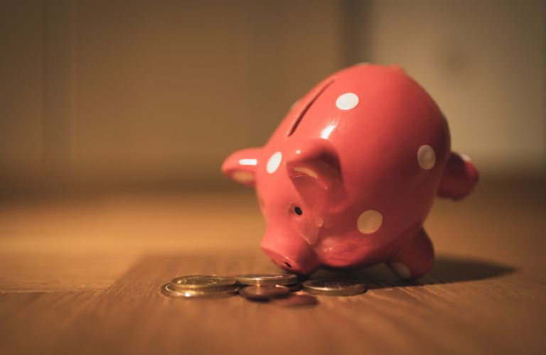 small piggy bank tilted forward as if to smell some coins lying on a wood table