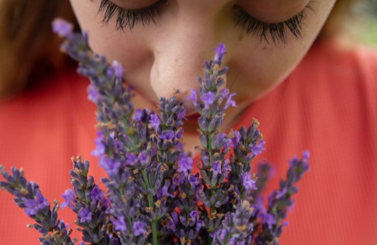 person in pink shirt smelling purple lilac flowers close up eyes closed