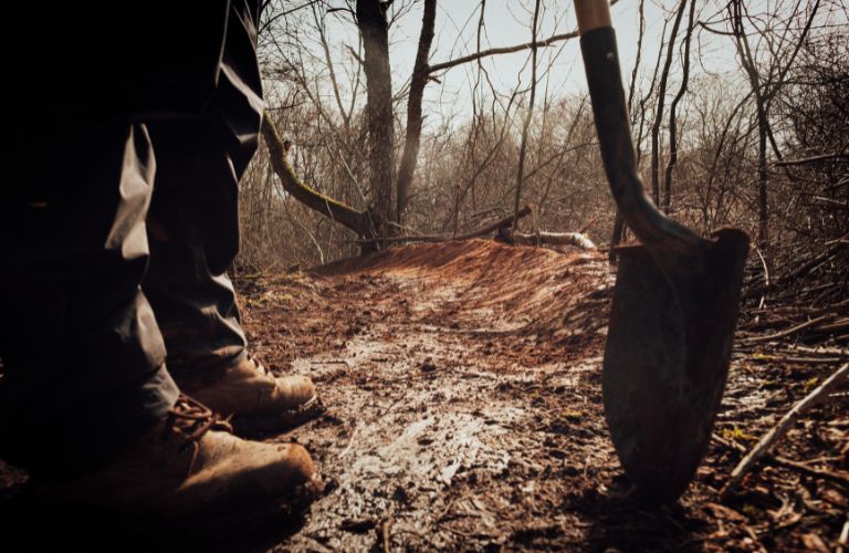 person with boots and shovel standing on forest path about to dig a hole
