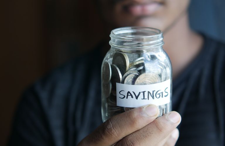 person holding up a glass jar labeled with the word savings and filled with coins of different denominations
