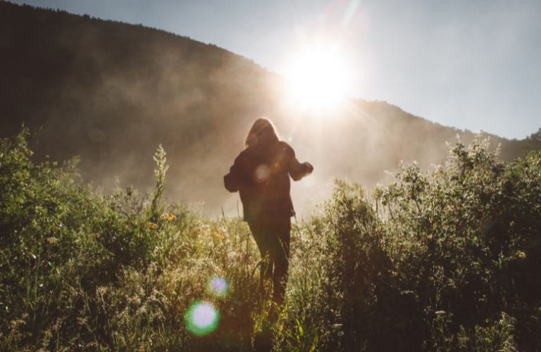 person walking through tall grasses and brush in the shadow of a hill