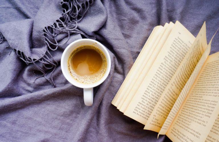 half drank cup of coffee next to an open book on a purple blanket